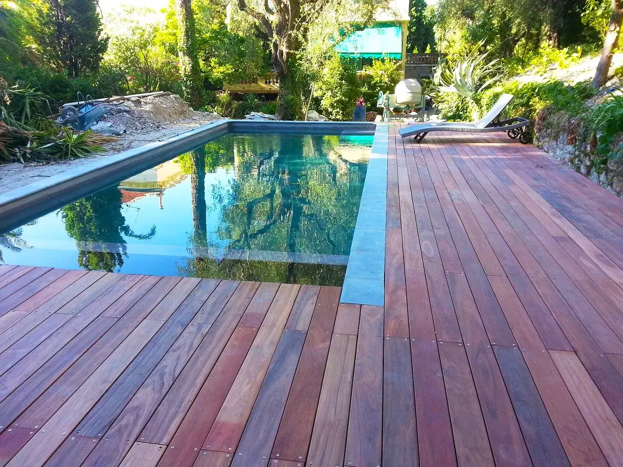 Terrasse en padouk vieillissant, reflétant le ciel et les arbres dans la piscine. Un transat attend au soleil.
