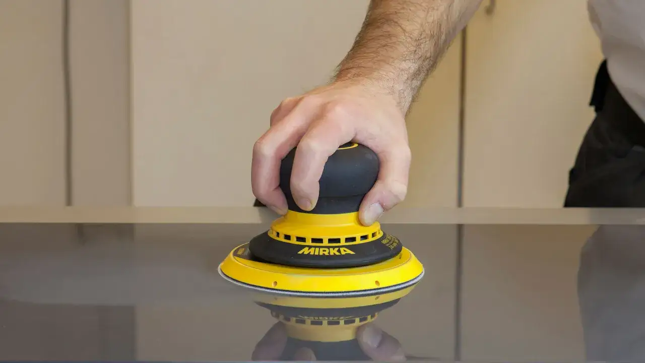 Main d'un homme utilisant une ponceuse Mirka jaune sur une surface sombre. Apprenez comment utiliser une ponceuse pour un fini parfait.