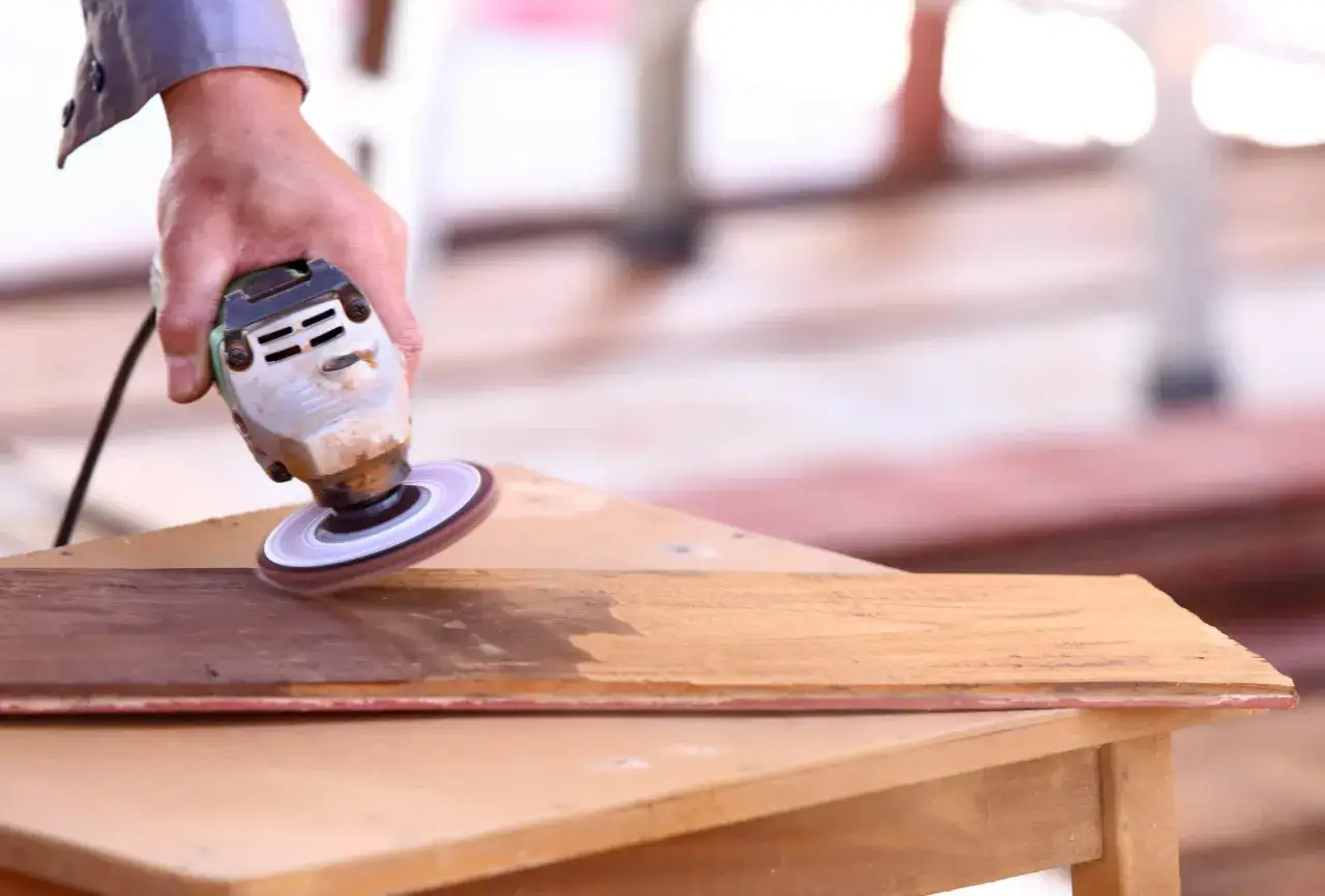 Main d'un artisan utilisant une ponceuse pour enlever une tache de peinture incrust&eacute;e sur du bois.