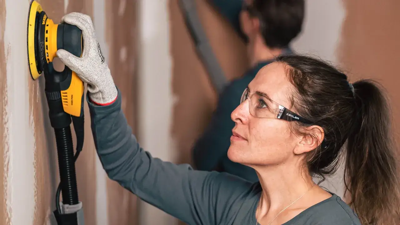 Une femme utilise une ponceuse pour préparer un mur avant de le peindre. Elle porte des lunettes de protection et des gants.