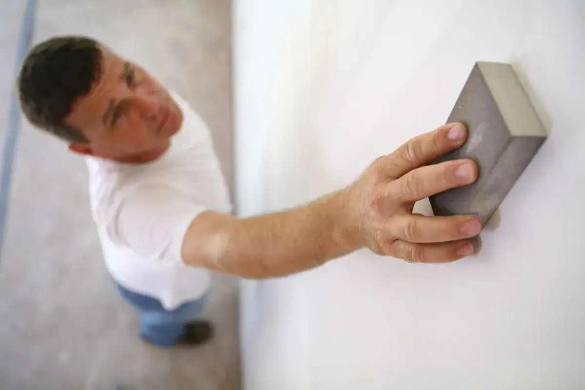 Un homme ponce un mur blanc avec une cale &agrave; poncer. Il pr&eacute;pare la surface, se demandant peut-on enduire sur de la peinture ?