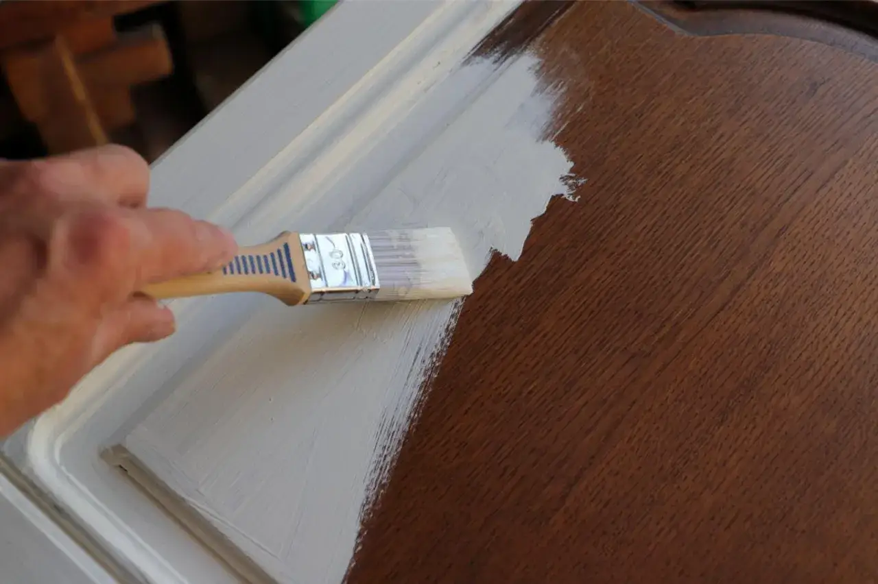 Une main applique de la peinture blanche sur un meuble en bois verni. La transformation commence, peindre sur du vernis pour un nouveau look.