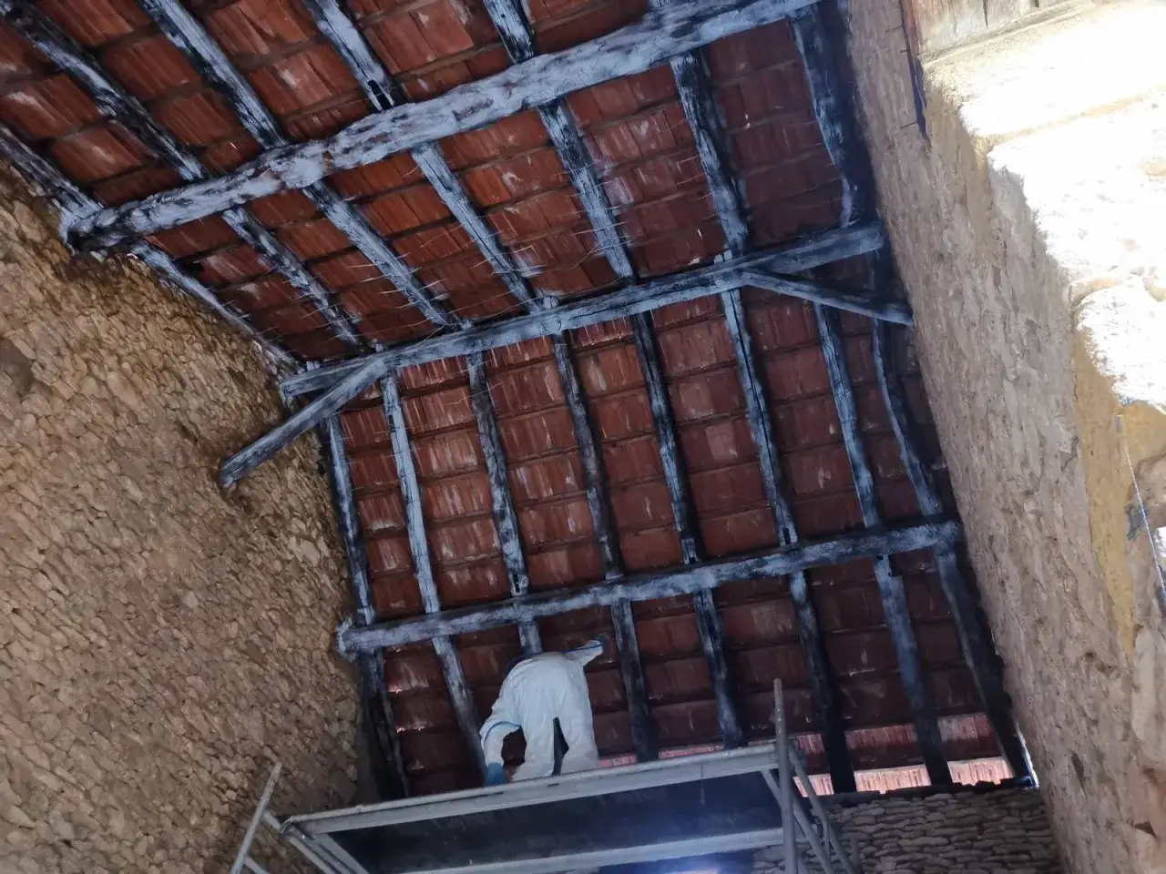 Une personne en combinaison blanche travaille sur un &eacute;chafaudage sous une charpente apparente bois et des tuiles rouges.