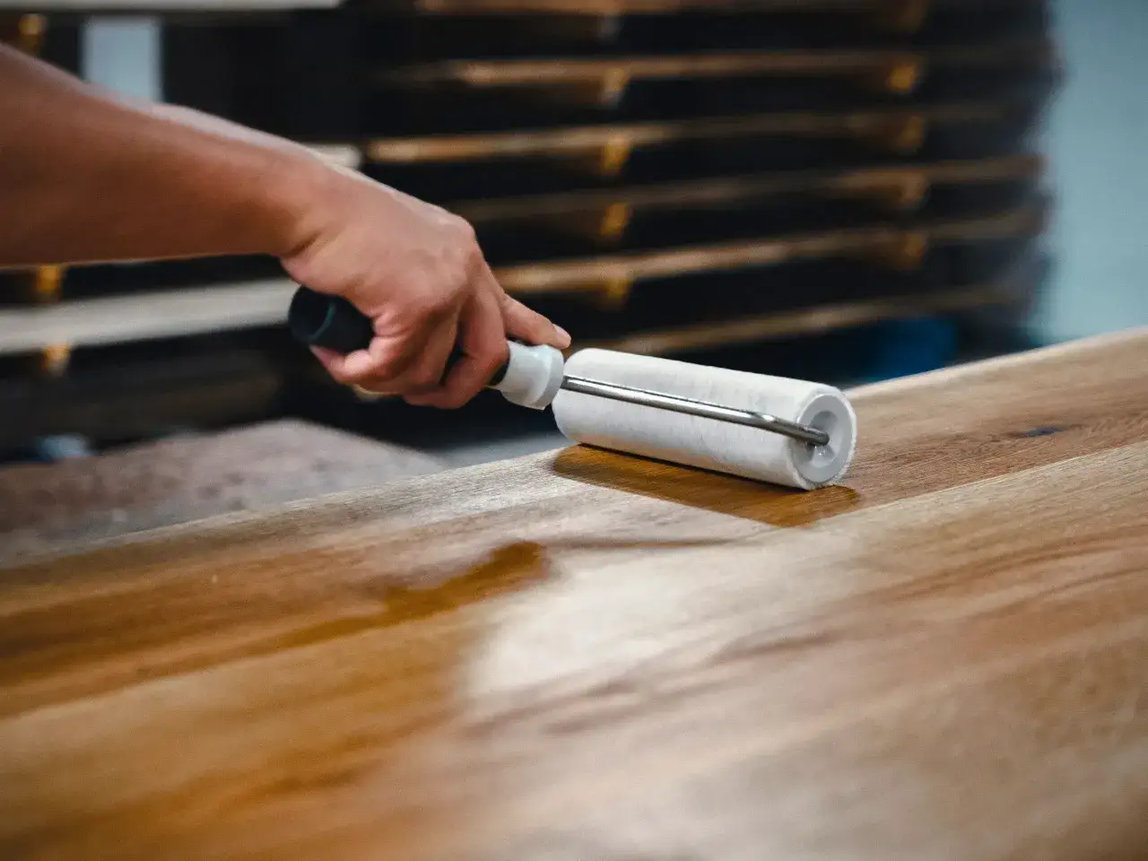 Main d'artisan appliquant une couche de vernis sur un plan de travail en bois avec un rouleau. Le bois est clair et présente des veines marquées.