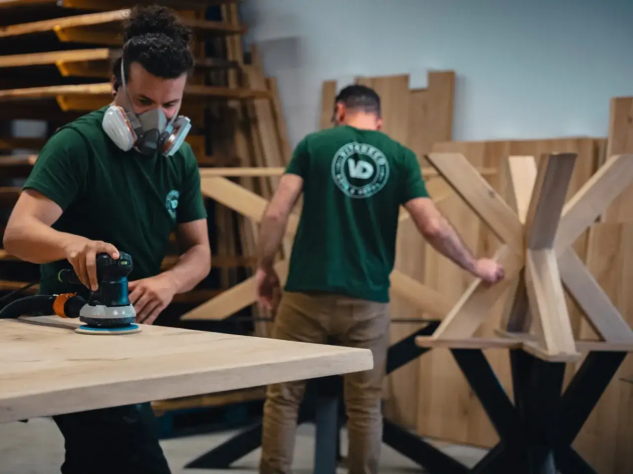 Artisan masqué en train de poncer une table en bois avec une ponceuse orbitale. Un autre artisan travaille sur un pied de table en bois en arrière-plan.