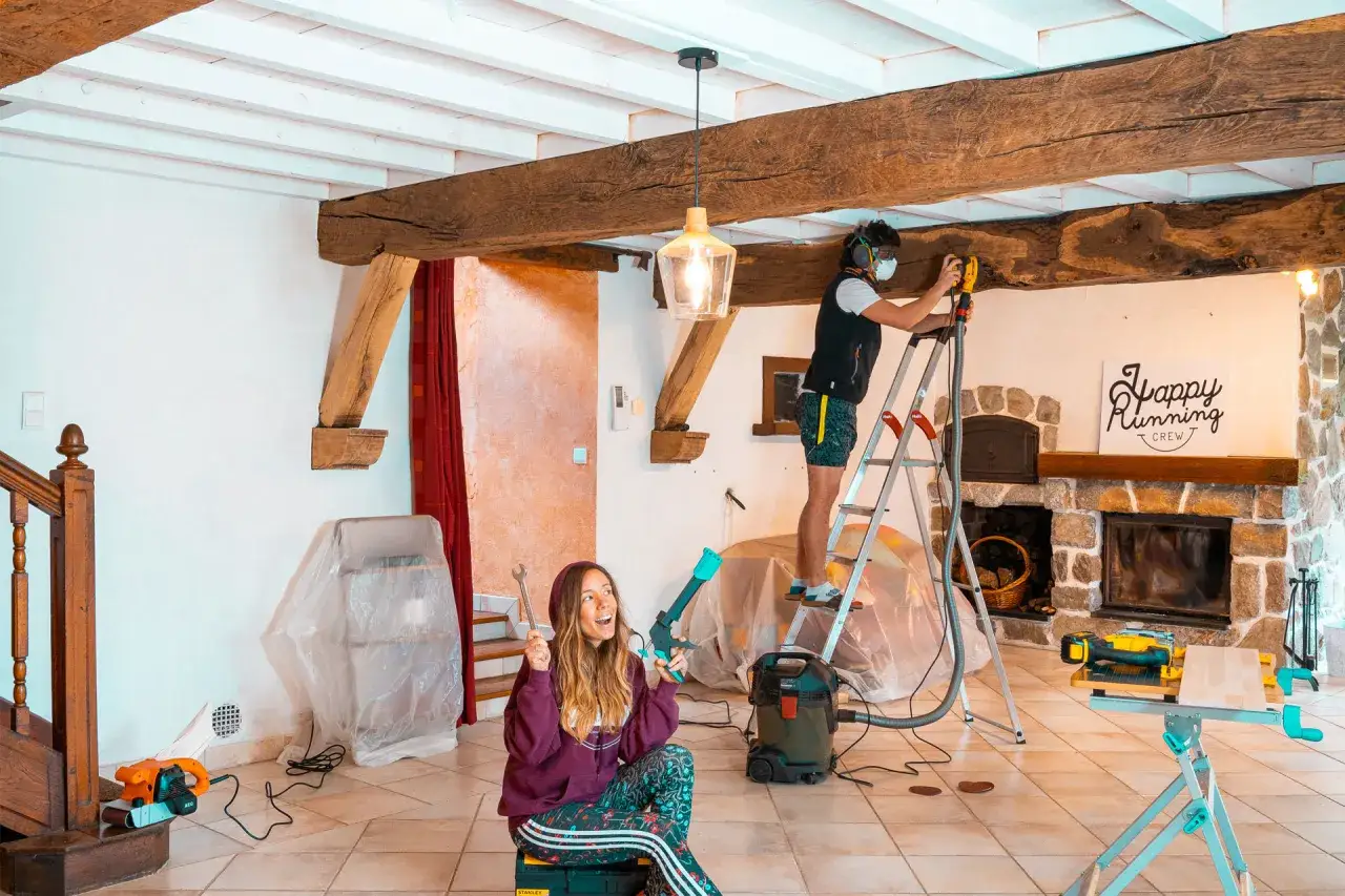 Un homme ponce une poutre en bois sur un escabeau, tandis qu'une femme tient des outils.