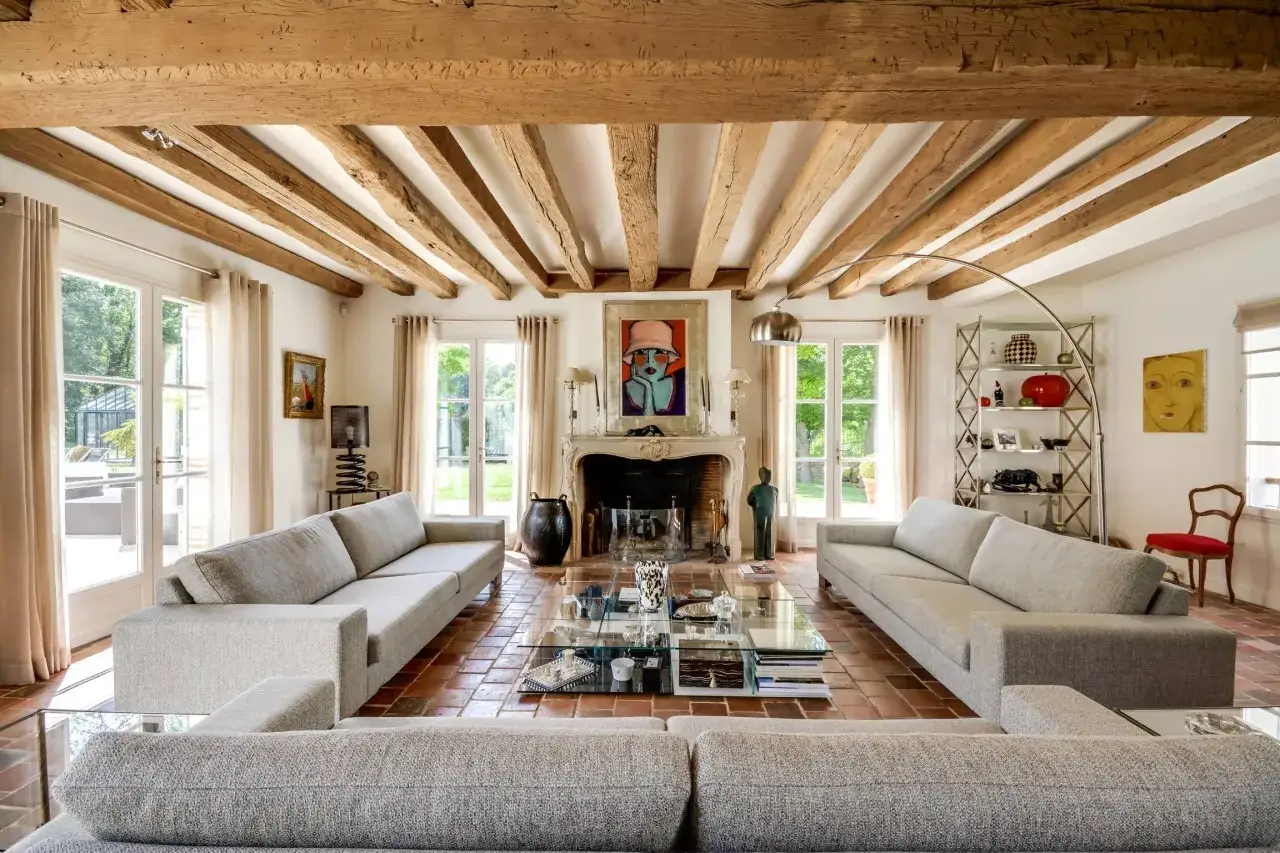 Salon lumineux avec poutres apparentes, un traitement bois brut intérieur qui souligne le caractère rustique. Canapés gris, cheminée et sol en terre cuite.
