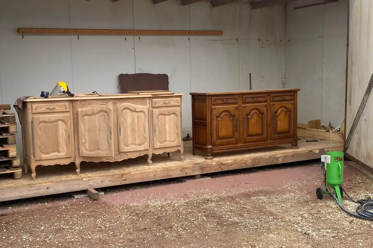 Deux commodes, l'une brute et l'autre vernie, attendent de nettoyer un meuble ancien. Un équipement de sablage vert est prêt à l'emploi.