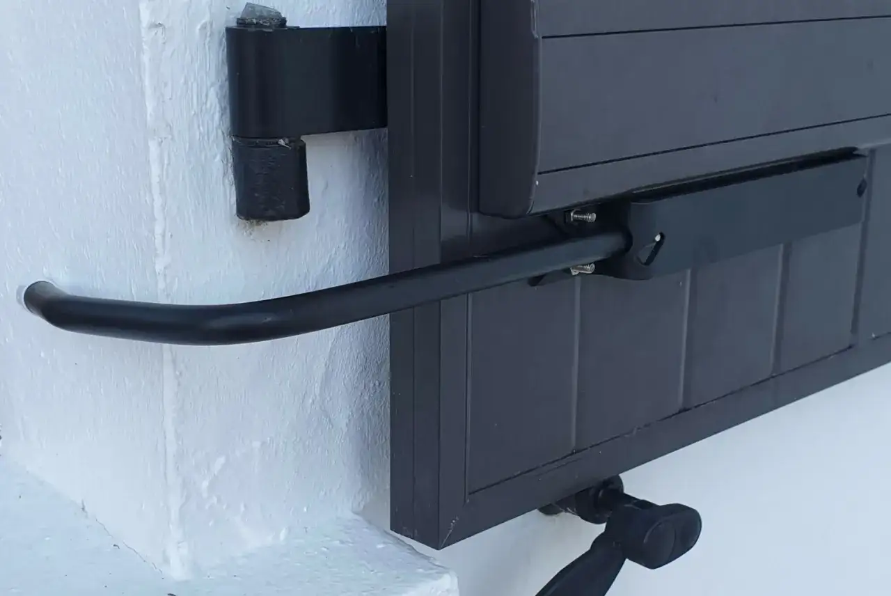 Un bras métallique noir, partie d'un arrêt de volets, est fixé à un volet gris sur un mur blanc.