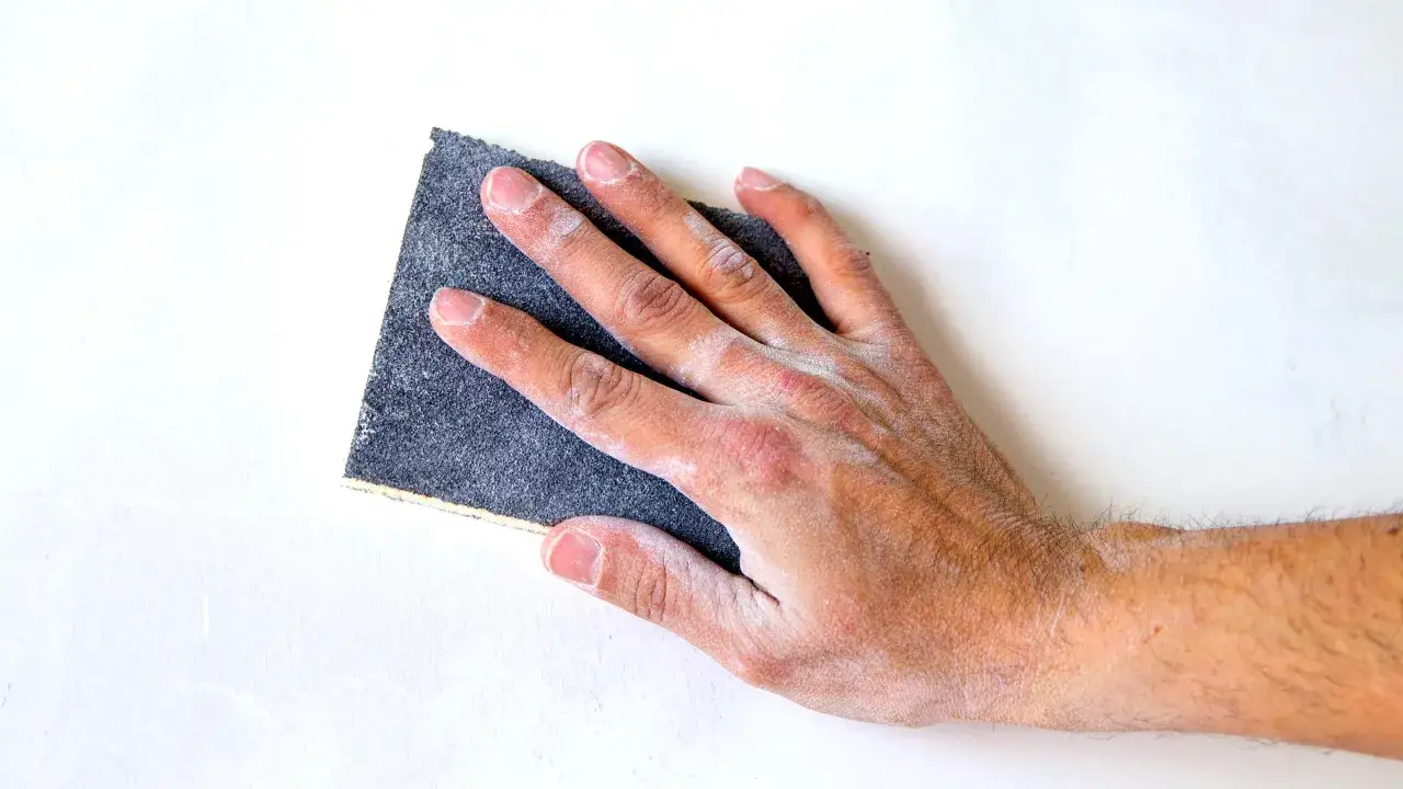 Main d'une personne utilisant du papier de verre pour poncer un mur en pl&acirc;tre.