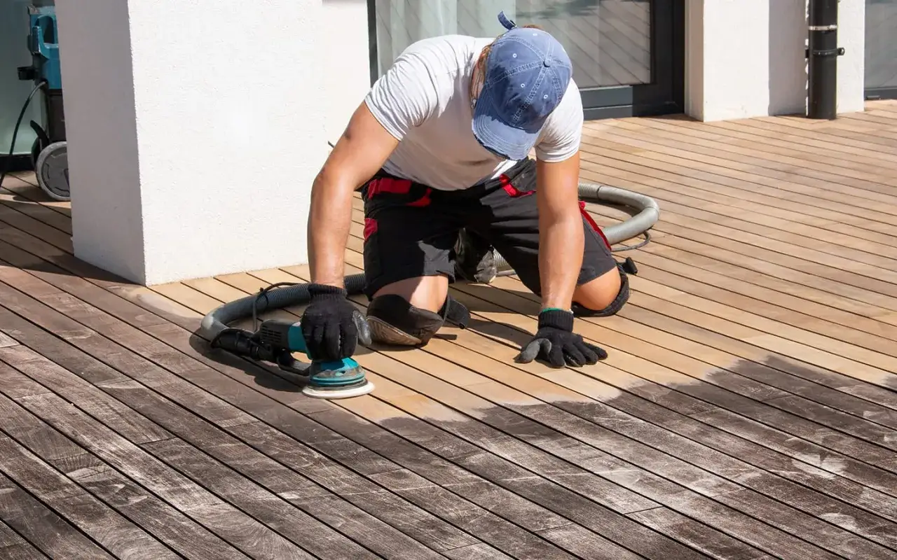 Un homme agenouillé ponce une terrasse en bois, montrant le processus d'entretien de terrasse bois.