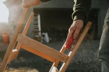 Main d'artisan ponçant un meuble en bois, préparant la surface pour enlever peinture sur bois acétone. Poussière de bois en suspension.