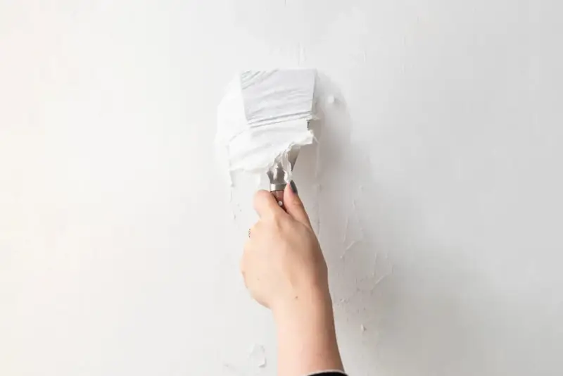 Une main utilise un couteau à mastic pour enlever de la vieille peinture écaillée d'un mur blanc, une étape essentielle pour savoir comment enlever la colle sur un mur avant de peindre.