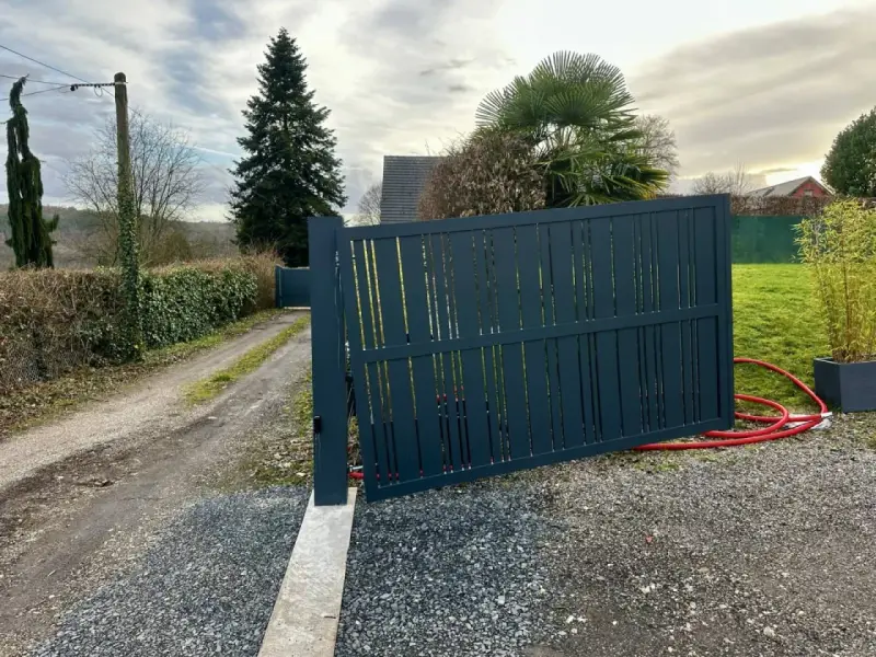 Portail en pente avec gonds sur mesure. Le portail gris foncé est ouvert, révélant un chemin de gravier et une allée bordée de haies.